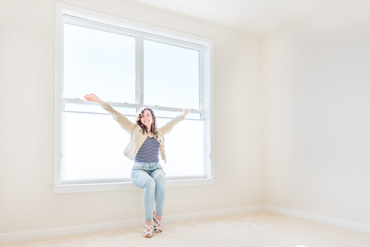 One Young Woman Sitting On Windowsill In White, Bright Empty, Clean Room On Carpet Of Modern New House, Home, Apartment Looking By Sunny Large Window With Blinds, Smiling Happy