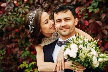 Handsome groom hugs radiant bride from behind while they stand before the wall of ivy