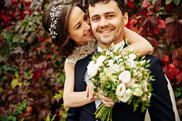 Handsome groom hugs radiant bride from behind while they stand before the wall of ivy