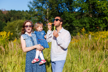 Young mother and father blowing soap bubbles and their little daughter trying to catch them. Funny kid playing outdoors. Concept on family holidays and walking at nature, parenting