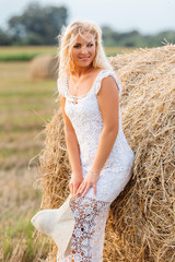 Naklejka na ściany i meble Beautiful tanned girl in white lace dress is standing near haystack at sunset in the summer. Happy, smiling woman in straw hat having fun, posing at the field. Concept of countryside lifestyle.