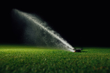 automatic lawn sprinkler spraying water over golf course green grass at night - obrazy, fototapety, plakaty