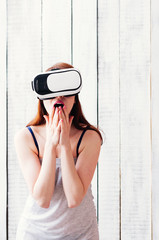 A girl in VR glasses, excited, holding her hands at face, white wooden background