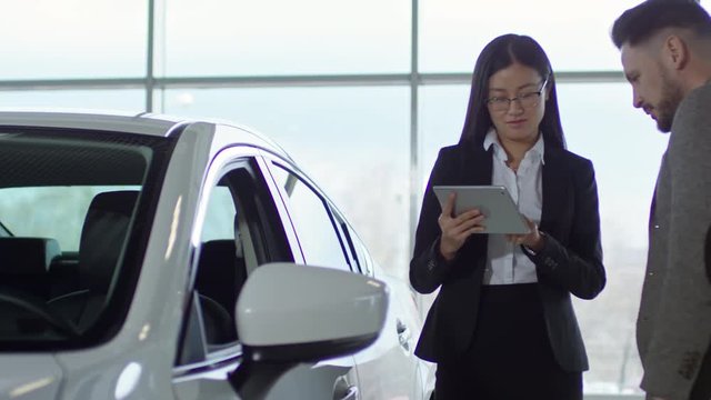 PAN of Asian saleswoman in glasses typing on tablet and talking with male client buying car at dealership