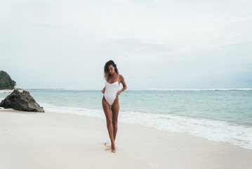 amazing girl in a white swimsuit with a beautiful sports body walking and posing on a white sand beach. A tanned young woman with curly hair is resting and sunbathing. Charming model posing near the