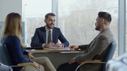 PAN of cheerful professional auto salesman in suit sitting at his desk and talking with couple signing purchase agreement, then giving them keys from new car and shaking hands