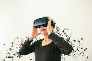 An elderly woman in virtual reality glasses is scattered by pixels. Conceptual photography with visual effects with an elderly person using modern technology.