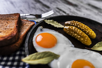 Fried eggs in a frying pan. Rye toast and pickled cucumber next to fried eggs.