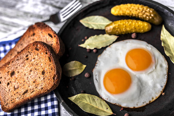 Fried eggs in a frying pan. Rye toast and pickled cucumber next to fried eggs.
