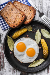 Fried eggs in a frying pan. Rye toast and pickled cucumber next to fried eggs.