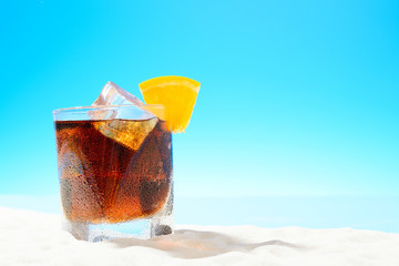 Refreshing Cola with ice and a slice of orange in a glass on the sandy coast