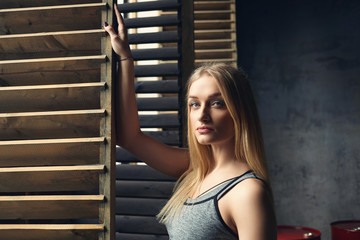 Portrait of beautiful blonde female with perfect slim body posing indoors in sportswear holding hand on window casement and looking at camera with serious confident expression on her pretty face