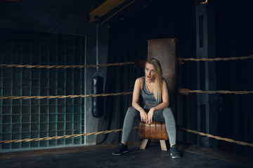 Obraz premium Indoor shot of gorgeous trendy looking young female model with loose dyed hair, natural make up and slim beautiful body sitting in the corner of boxing ring, posing in fashionable top and leggings