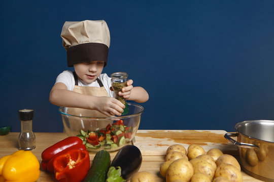 Adorable male child helping mother cooking salad for whole family, posing isolated at blank wall, holding glass bottle, adding spices into bowl, having focused look, vegetables and casserole on table