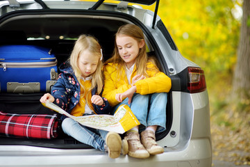 Two adorable girls sitting in a car trunk before going on vacations with their parents. Two kids looking forward for a road trip or travel. Autumn break at school.