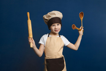 Fototapeta premium Children, cooking, cuisine and food concept. Picture of funny little boy wearing chef uniform standing at blank wall, holding rolling pin in one hand and two wooden spoons in other, smiling at camera