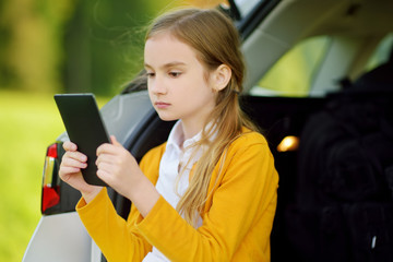 Adorable little girl ready to go on vacations with her parents. Kid sitting in a car trunk and reading her ebook.