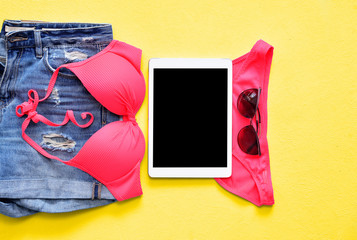 Female summer bikini swimsuit and accessories on blue with tablet and sunglasses.
