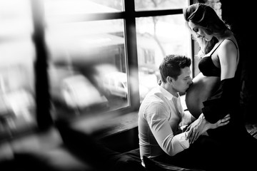 Black and white photo on which husband sensually kisses the belly of his beautiful pregnant wife on the bed near the window