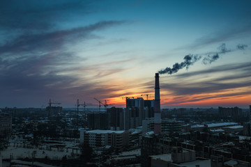 Panorama of city sunset and silhouettes of cranes, high-rise buildings and construction site with smoke