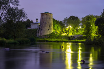 Old stone tower of medieval fortress and small church reflecting in river at night. Pskov fortifications, Russia.