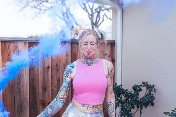 Wide portrait of blonde caucasian woman with tattoos using blue smoke bomb in an urban location