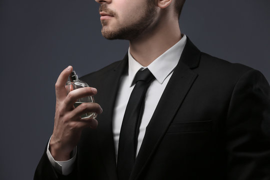 Handsome man in suit using perfume on dark background, closeup