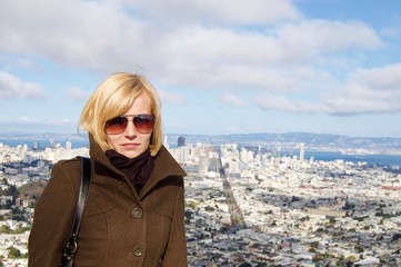 Beautiful blonde female tourist visiting the San Francisco Twin Peaks overlooking the city's scenic skyline with numerous buildings and landmarks in the background - California, USA