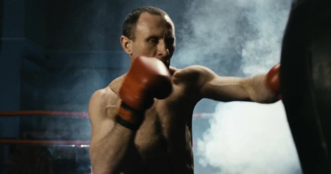 Serious boxer in gloves training Thai box while attacking punching bag in smoke on ring.