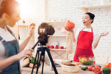 Two culinary bloggers hold up pumpkin with one girl behind camera.
