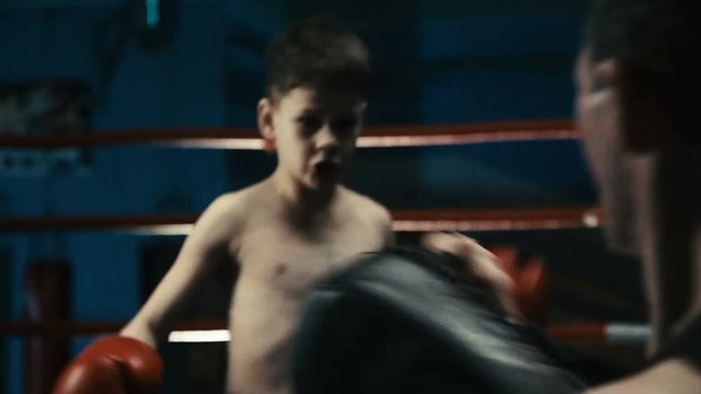 Back view of professional coach holding punching bag and working out with boy teaching boxing attacks.