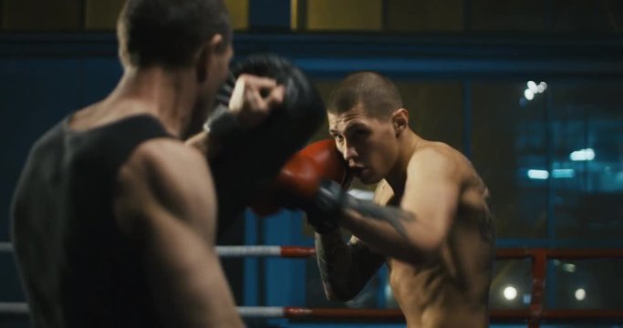 Handheld slowmotion 4K shot of shirtless muscular boxer with tattooed body working out on ring in gym attacking coach with punching bags.