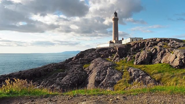 White and grey clouds passing over the historic Ardnamurchan Lighthouse in Western Scotland on a summer day. Black rocks, blue ocean and distant islands are visible. Low angle shot from the ground.