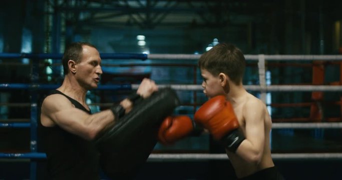 Side view of professional coach holding punching bag and working out with boy teaching boxing attacks.