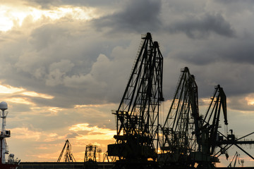 Obraz premium Silhouette of port cranes against a dramatic sky at sunset.