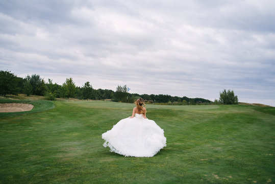 Full length body portrait of beautiful bride in fashion white wedding dress with feathers running away through green golf course, back view. Runaway bride, copy space