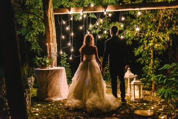 Night wedding ceremony with candles, lanterns and lamps on tree. Bride and groom holding hands on background of baulb lights, back view. Beautiful young couple standing under a tree at night - Powered by Adobe