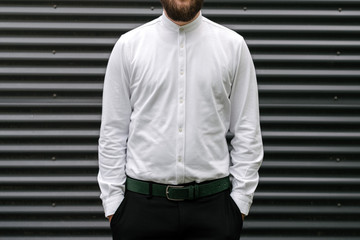 Confident young bearded man in white shirt keeping arms in pockets while standing against grey metal background