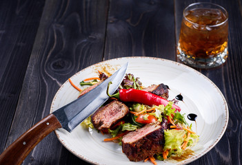 Veal salad with lettuce, carrot, tomato and pepper on white plate on dark wooden background, copy space. Restaurant food, close up. Meat salad and glass of whiskey