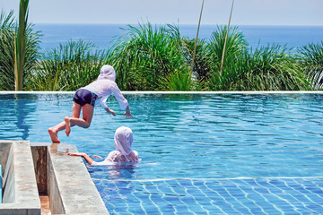 Children play in pool in clothes for protection from ultraviolet rays. Outdoor pool on background  palm and sea. Back view.