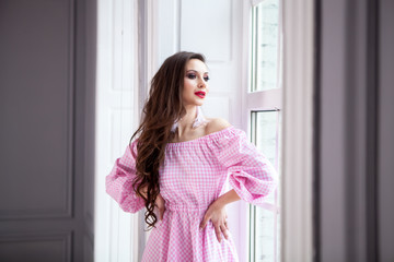 young beautiful girl stands at the big window in the floor and looks out into the distance. Dressed in a light checkered pink dress, long hair, red lips