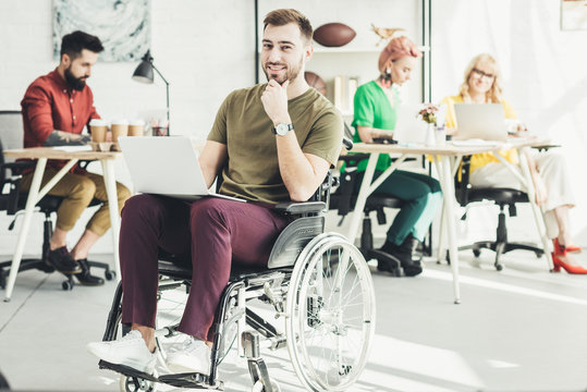 selective focus of smiling disabled businessman with laptop and colleagues working behind in office