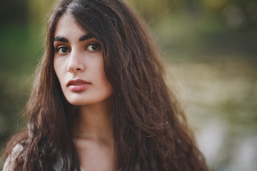 Fototapeta premium Lovely brooding brunette girl with long hair in white romantic dress stands on a wooden bridge in spring forest or park. The atmosphere of a warm sunny evening. The background is blurred.