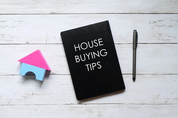 Top view of house model,pen and notebook written with 'HOUSE BUYING TIPS' on white wooden background.
