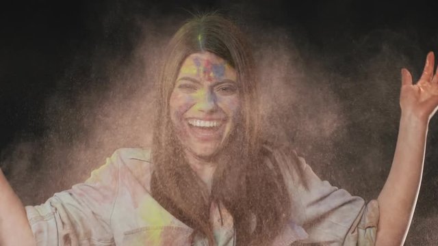 Portrait of ecstatic woman making splash with colorful powder under bright paints on holi spring festival, isolated over dark gray background slow motion. Indian culture and tradition