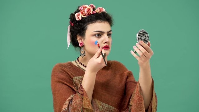 Portrait of woman style icon and famous celebrity Frida Kahlo painting her face with brush and making art or performance, isolated over green background. Stylization concept