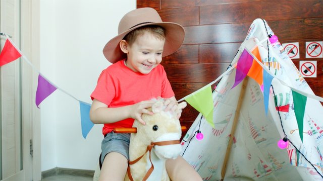 Little happy boy wearing cowboy hat is riding a toy horse in the playroom