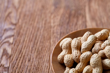 Unpeeled peanuts in wooden plate over rustic wooden background closeup, selective focus.