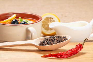 Wooden spoon with black peppercorn, dried red pepper on cutting board and ceramic soup bowl with saltwort, sauceboat and cut lemon