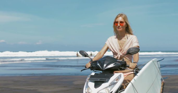 Close-up of young blonde girl driving motorbike with surfboard on the beach near the water. 4k video shooting in motion in front